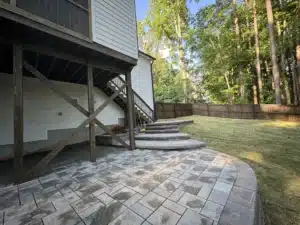 A view of a newly constructed patio featuring Techo Bloc Champegne Grey pavers with a slate finish, nestled under a house deck. The patio is bordered by a sturdy 4 ft Versalock retaining wall and curved steps, surrounded by lush Zenith zoysia grass sod and tall trees, showcasing the hardscaping transformation.