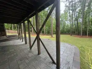 A close-up view under the deck showing the intricate layout of Techo Bloc Champegne Grey paver patio, with a sturdy 4 ft retaining wall made of Versalock blocks.