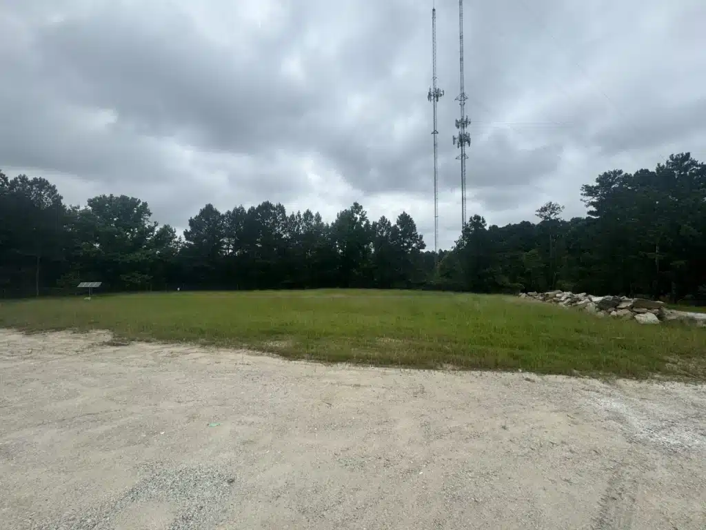 A wide shot of a Durham hydroseeding project at a glass plant site in Durham, NC, featuring a grassy area with two tall communication towers under a cloudy sky, surrounded by trees and a gravel foreground with a rock barrier on the right.