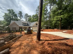 A modern home with a gray roof and white siding under construction, surrounded by tall trees, with a concrete driveway and grading equipment visible on a dirt lot.