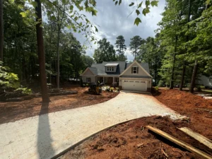 A modern home with a gray roof and white siding, surrounded by tall trees, featuring ongoing grading work with a concrete driveway and grading equipment on red dirt.