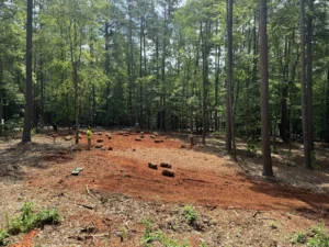 A section of red dirt yard with scattered bales of hydroseeded Kentucky 31 Fescue, workers in the background, and tall trees framing a modern home.