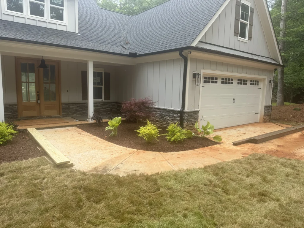 A landscaped bed with Designer Brown Mulch, featuring plants like Loropetalums and Japanese Maples, next to a concrete walkway leading to a house.