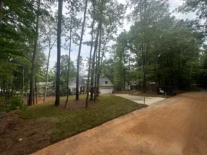 A newly poured concrete driveway curves toward a white house with a garage, flanked by freshly laid Fescue sod and surrounded by trees and red dirt.