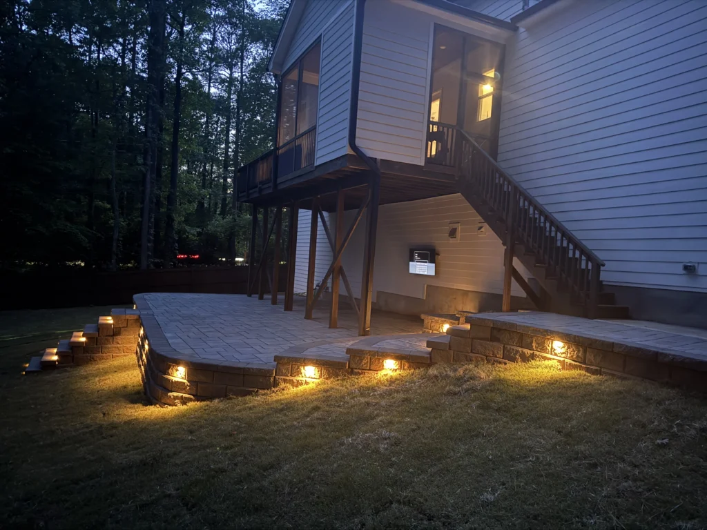 A beautifully lit backyard at dusk featuring a modern house with a deck, illuminated by warm outdoor lights along a newly constructed retaining wall and steps made with Versalock retaining wall blocks.
