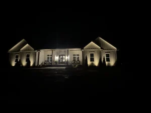 A night view of a well-lit house in Durham with outdoor lighting highlighting the facade, featuring a covered porch with chairs and potted plants, set against a dark sky.