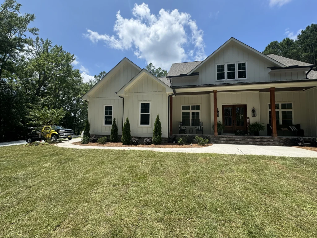 : A daytime view of a house in Durham with lush new St. Augustine sod covering the front yard, a driveway with a yellow vehicle, and neatly landscaped beds with small shrubs under a blue sky with clouds.