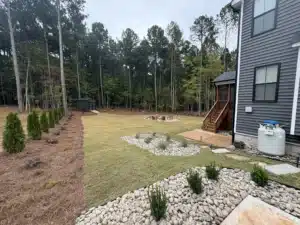Wide view of the completed backyard in Franklinton, NC, showing fresh Bermuda sod, defined plant beds with Delaware river rock and metal edging, a Techo-Bloc Valencia fire pit area with pea gravel, landscape lighting, and a variety of newly planted shrubs and trees against a wooded backdrop.